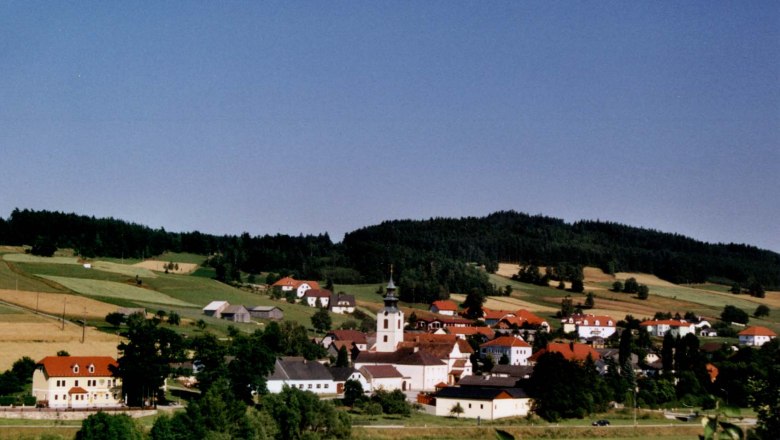 St. Martin, © Marktgemeinde St. Martin Panorama eines Dorfes mit Kirche und roten Dächern, umgeben von grünen Feldern und Wäldern.