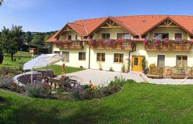 Fewo-Pöcher, © Walter Pöcher Ein großes gelbes Haus mit roten Dächern und Blumenkästen, umgeben von einem gepflegten Garten mit Sitzbereich und Sonnenschirm.