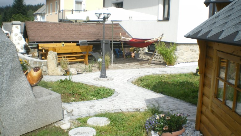 Gartenbereich, © Manfred Gill Garten mit Steinen, Holzhaus, Tisch, Hängematte und Sonnenschirm.