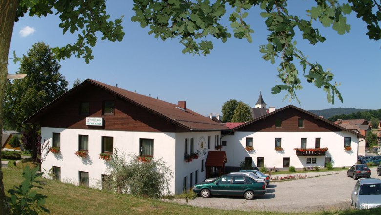 Landgasthof Binder, © Fam. Binder Ein Landgasthof mit weißen Wänden und braunem Dach, umgeben von Bäumen und Autos auf einem Parkplatz.
