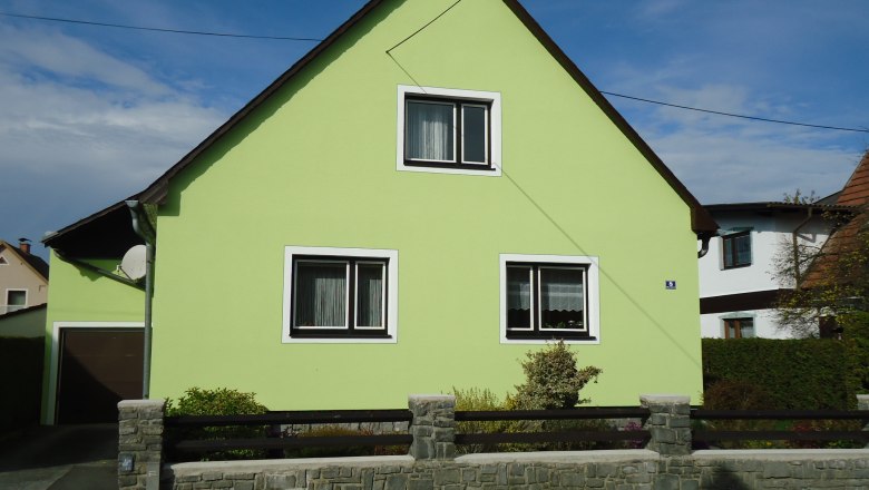 Ein hellgr&uuml;nes Haus mit Satteldach und zwei Fenstern, umgeben von einem niedrigen Steinzaun.