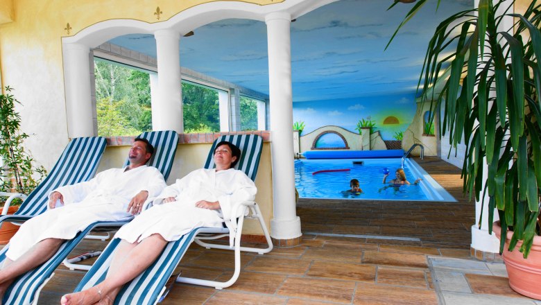 Zwei Personen entspannen auf Liegest&uuml;hlen im Wellnessbereich neben einem Innenpool.