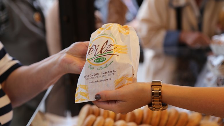 Zwei H&auml;nde &uuml;berreichen eine Papiert&uuml;te der B&auml;ckerei-Konditorei Pilz mit Geb&auml;ck im Hintergrund.