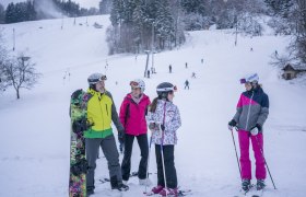 Gruppe von Skifahrern und Snowboardern im Schnee vor einer Skipiste mit Lift.
