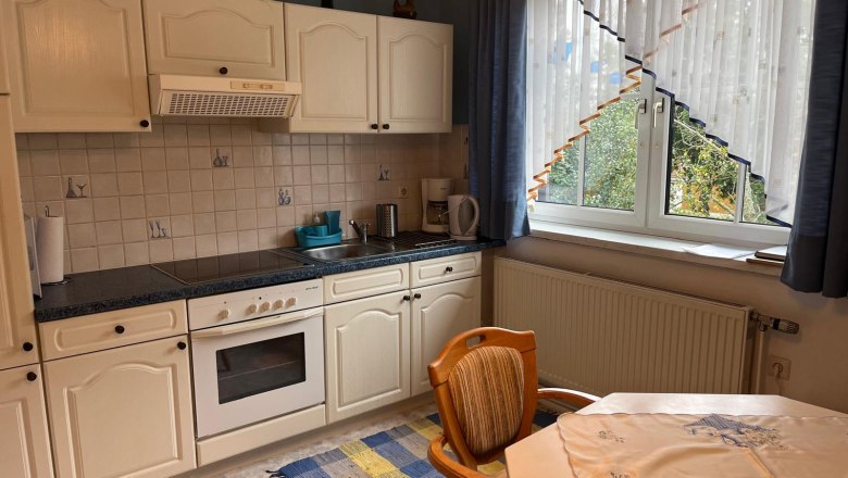 K&uuml;che mit wei&szlig;en Schr&auml;nken, Herd, Sp&uuml;le und Fenster mit Vorh&auml;ngen.