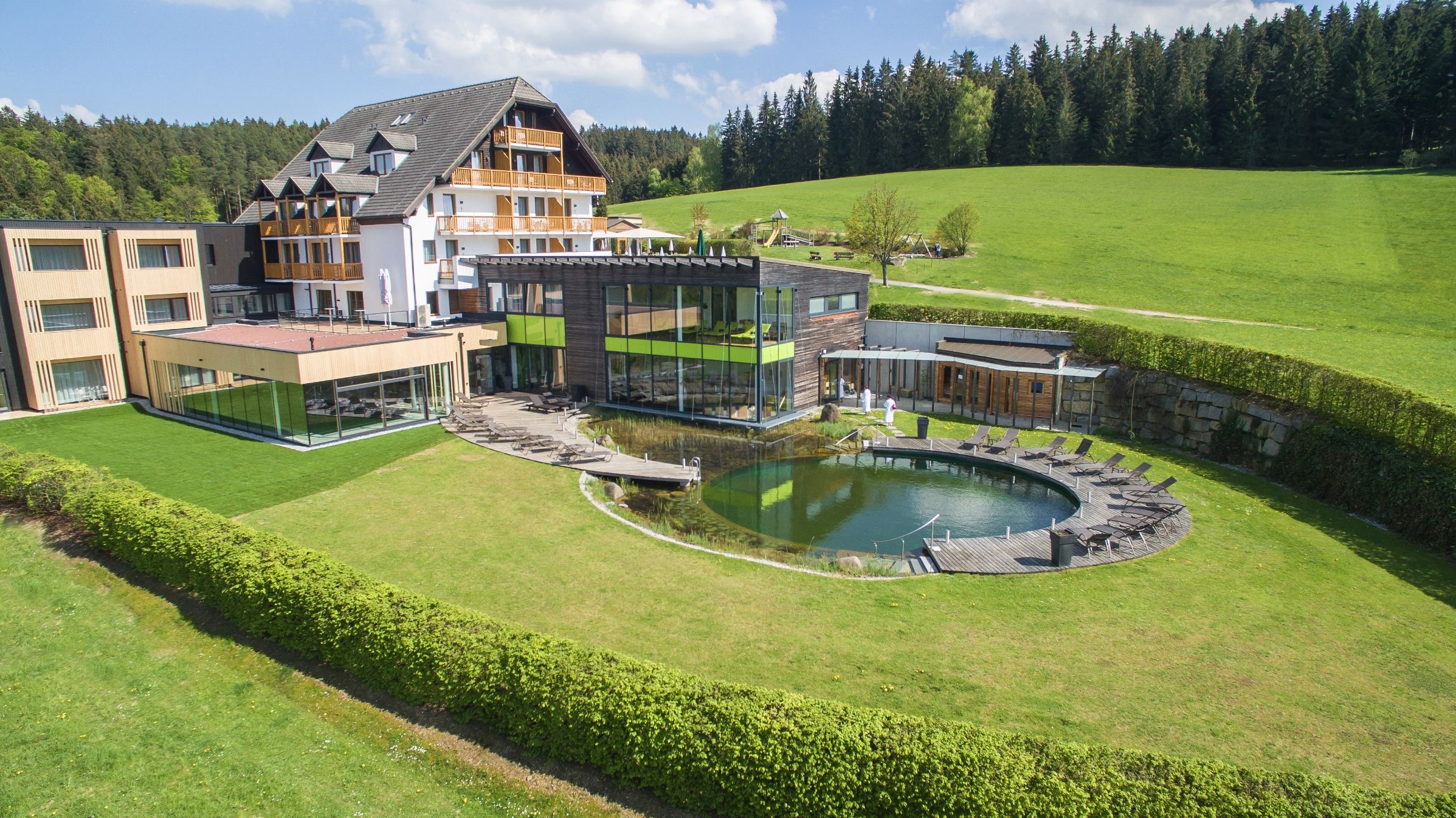 Hotel Schwarz Alm Zwettl mit Naturteich und Liegestühlen, umgeben von grüner Landschaft.