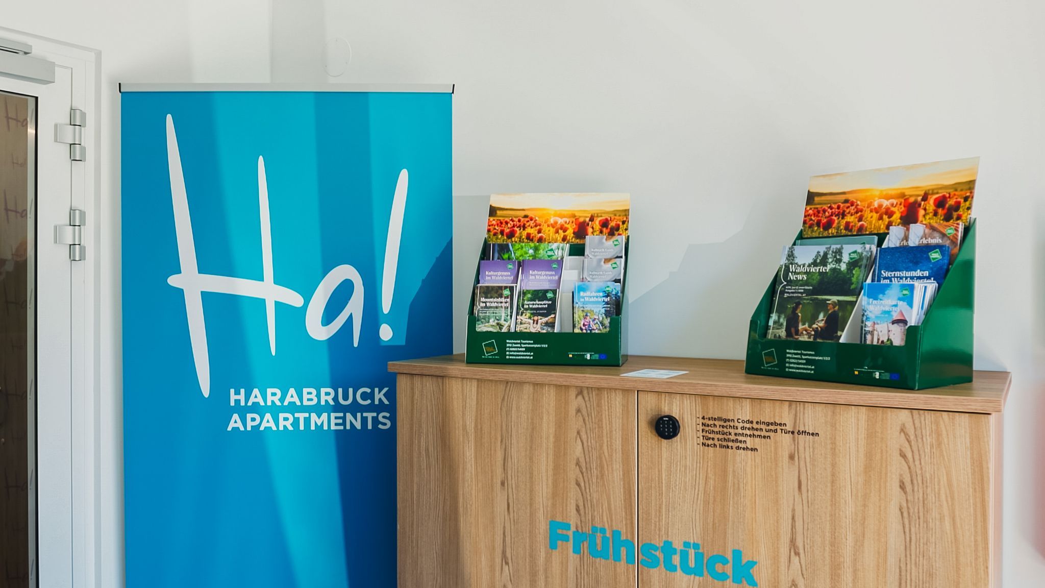 Ein Empfangsbereich mit einem blauen Banner der Harabruck Apartments und einem Prospektständer auf einem Holzschrank.