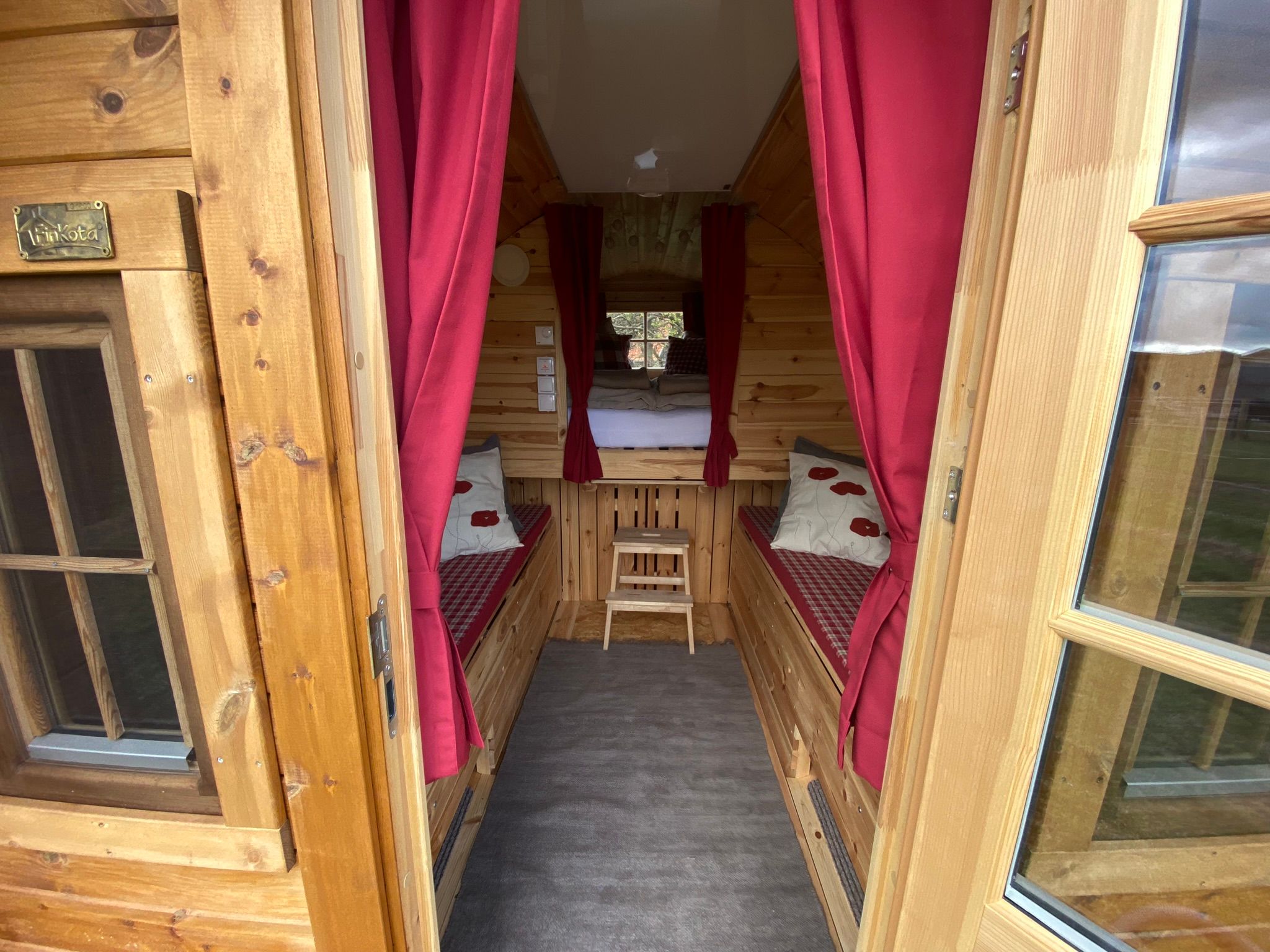 Innenansicht eines Holzfasses mit Bänken und einem Bett im Hintergrund.