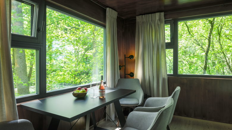Innenansicht eines modernen Baumhauses mit Tisch, St&uuml;hlen und gro&szlig;en Fenstern mit Blick ins Gr&uuml;ne.