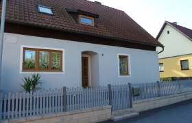 Ein Einfamilienhaus mit grauer Fassade, braunem Dach und Holzt&uuml;ren. Ein kleiner Zaun umgibt das Grundst&uuml;ck.