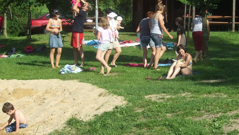 Menschen entspannen an einem Naturbadeteich mit Sonnenschirm und B&auml;umen.