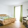 Helles Zimmer mit Holzbett, gr&uuml;n-wei&szlig; gestreifter Bettw&auml;sche, Tisch mit Vase und Pflanze, gr&uuml;ne Vorh&auml;nge.