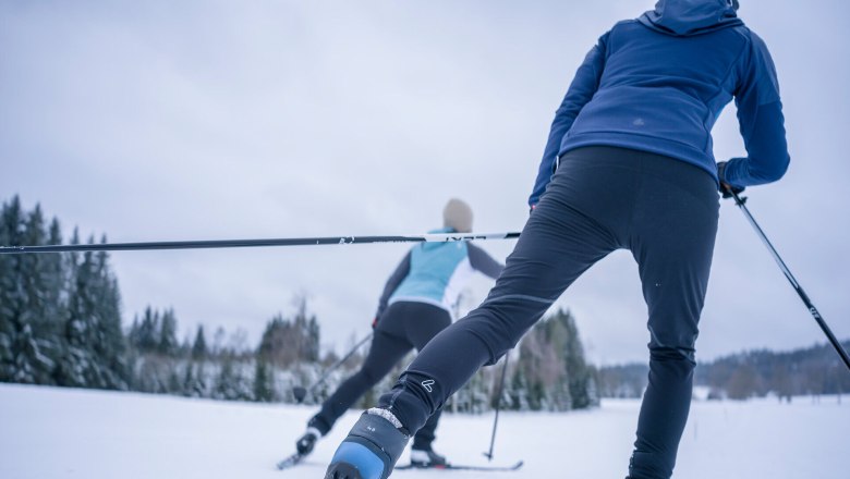 Zwei Personen beim Langlaufen auf einer verschneiten Strecke, umgeben von B&auml;umen.