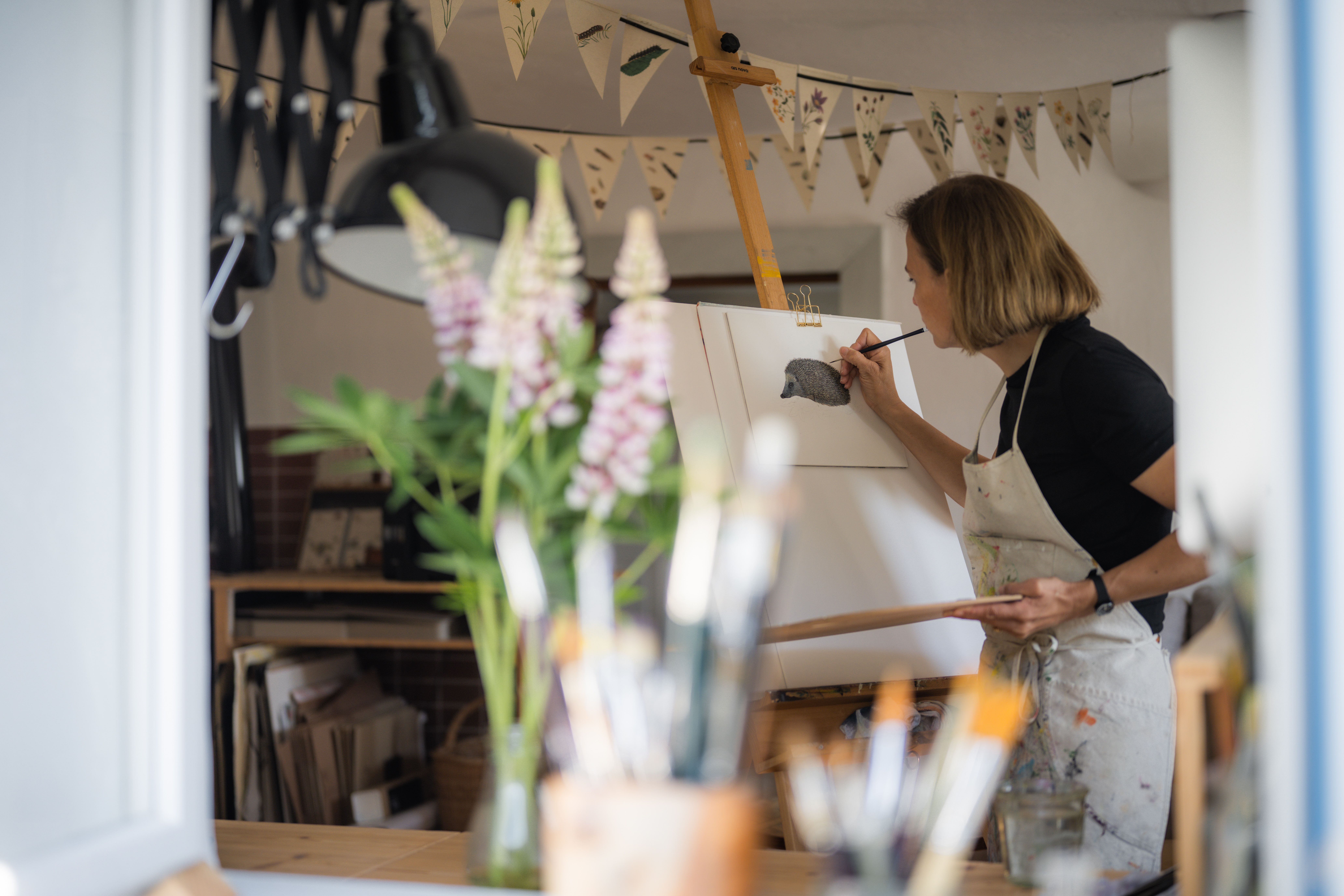 Eine Frau malt in einem Atelier an einer Staffelei ein Bild von einem Igel. Im Vordergrund sind Pinsel und Blumen zu sehen.
