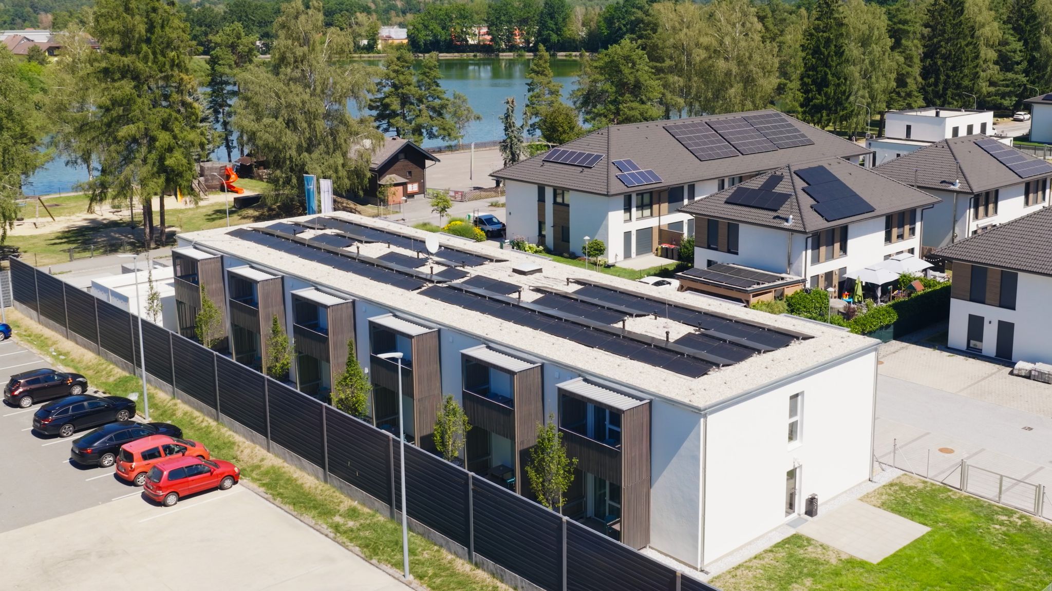 Luftaufnahme von modernen Gebäuden mit Solaranlagen auf den Dächern, umgeben von Bäumen und einem See im Hintergrund.