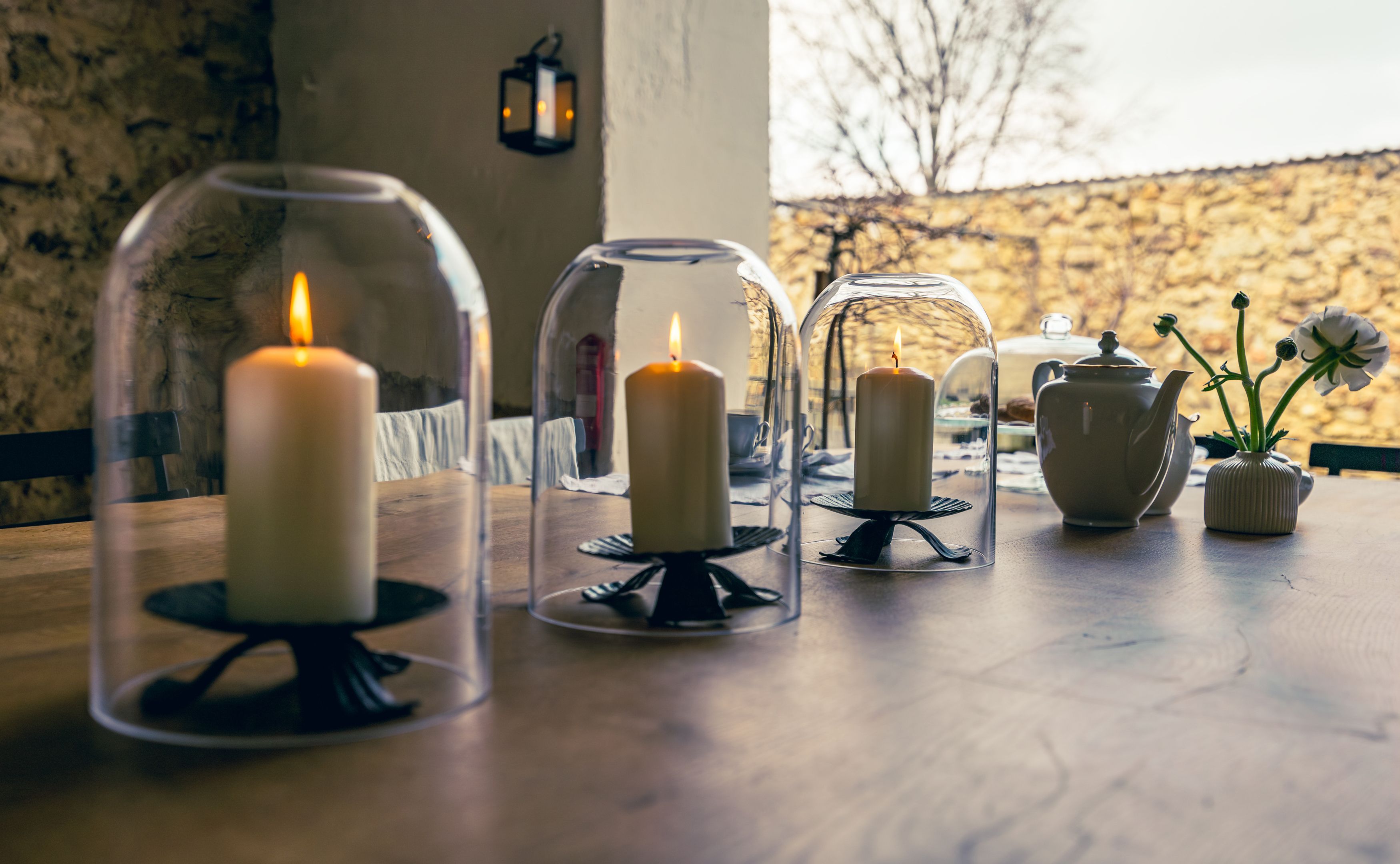 Drei brennende Kerzen unter Glasglocken auf einem Holztisch, daneben eine Teekanne und eine Vase mit Blumen.