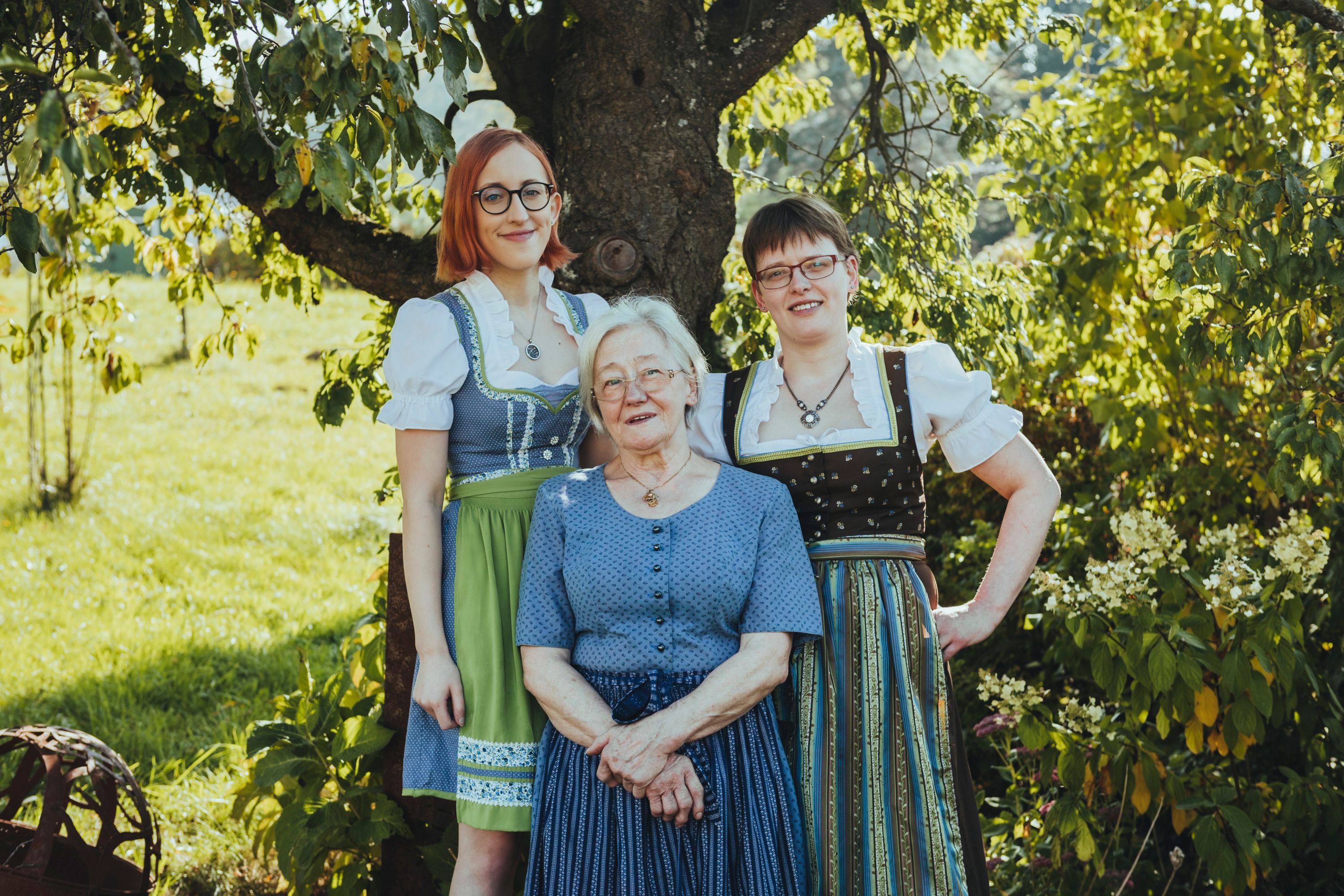 Drei Frauen in traditioneller Tracht stehen lächelnd unter einem Baum im Freien.