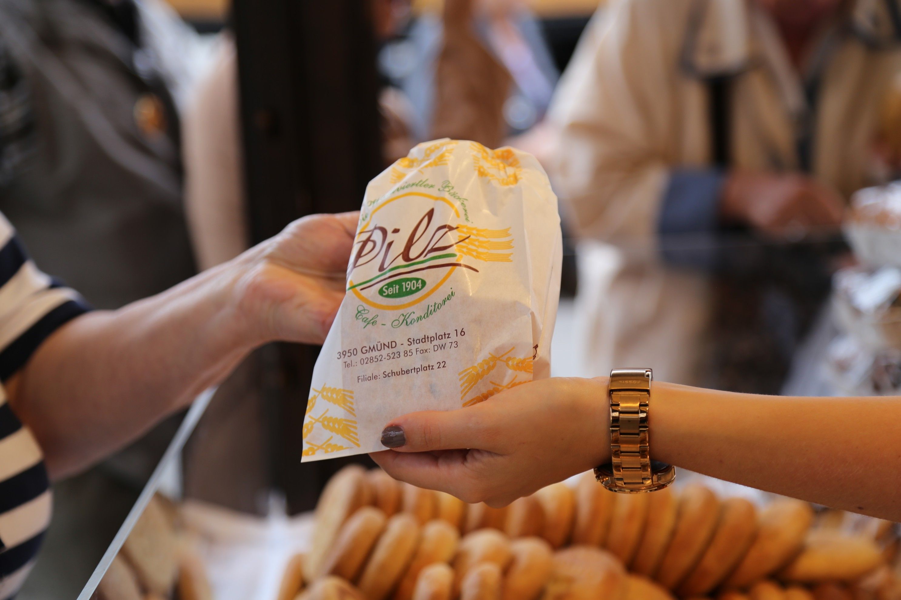 Zwei Hände überreichen eine Papiertüte der Bäckerei-Konditorei Pilz mit Gebäck im Hintergrund.