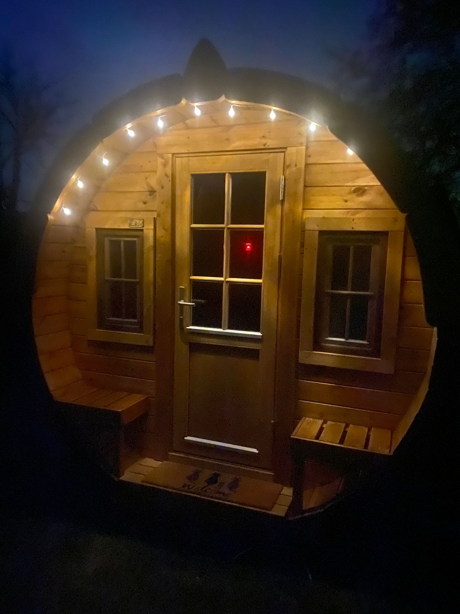 Ein kleines, rundes Holzhaus mit Lichterkette über der Tür, aufgenommen bei Nacht.