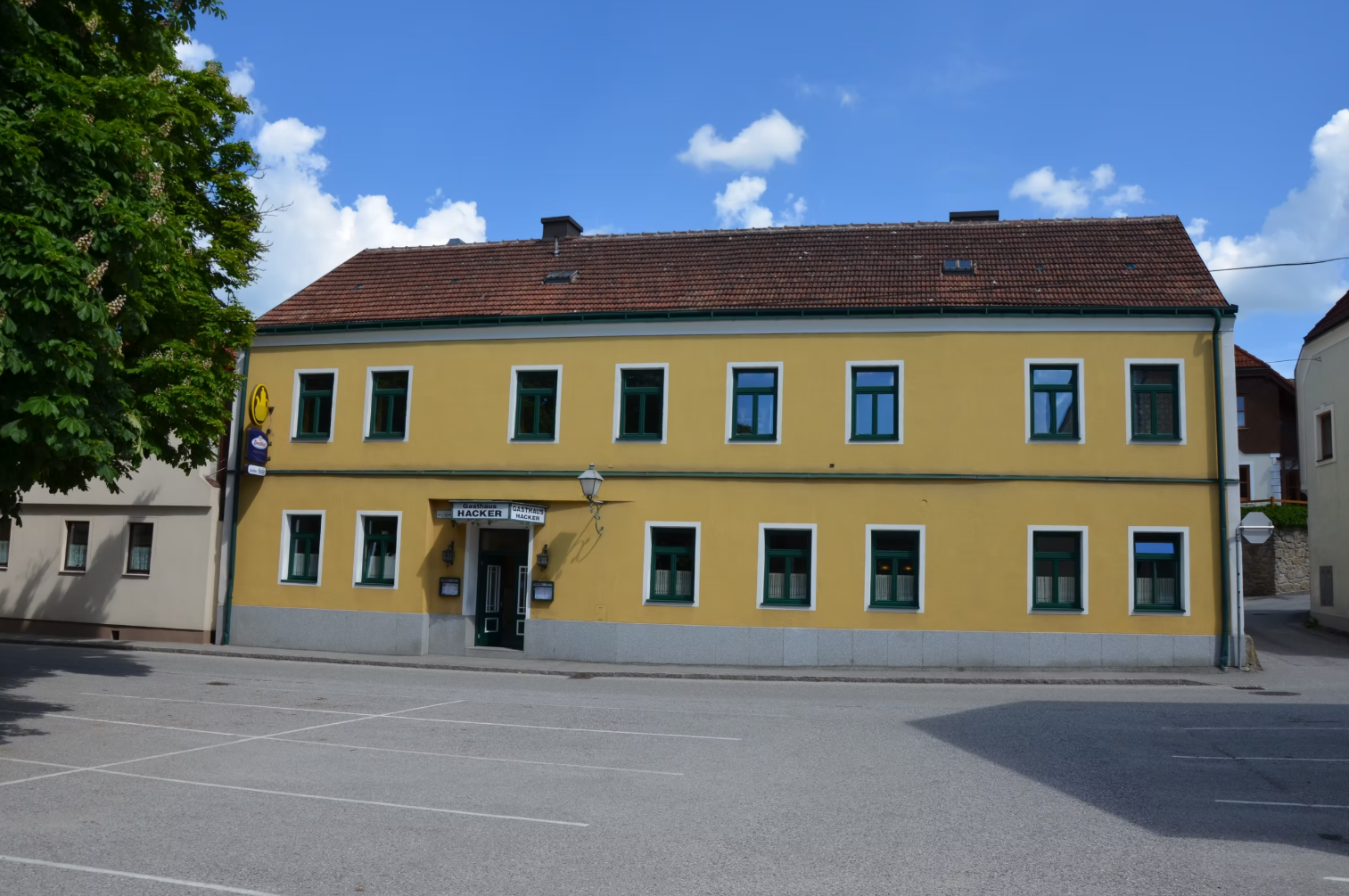 Gelbes zweistöckiges Gebäude mit grünen Fensterrahmen und einem Schild mit der Aufschrift 'Gasthaus Georg Hacker'.