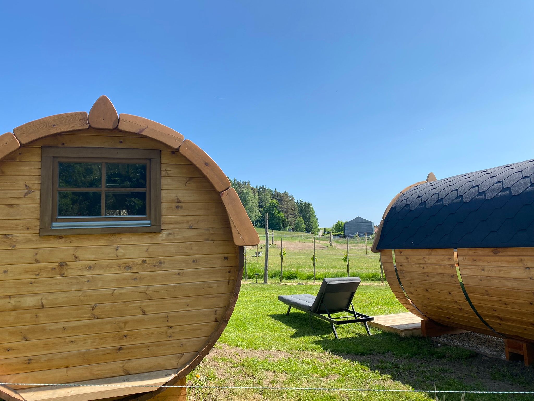 Zwei Schlaffässer aus Holz mit einer Liege auf einer Wiese bei sonnigem Wetter.