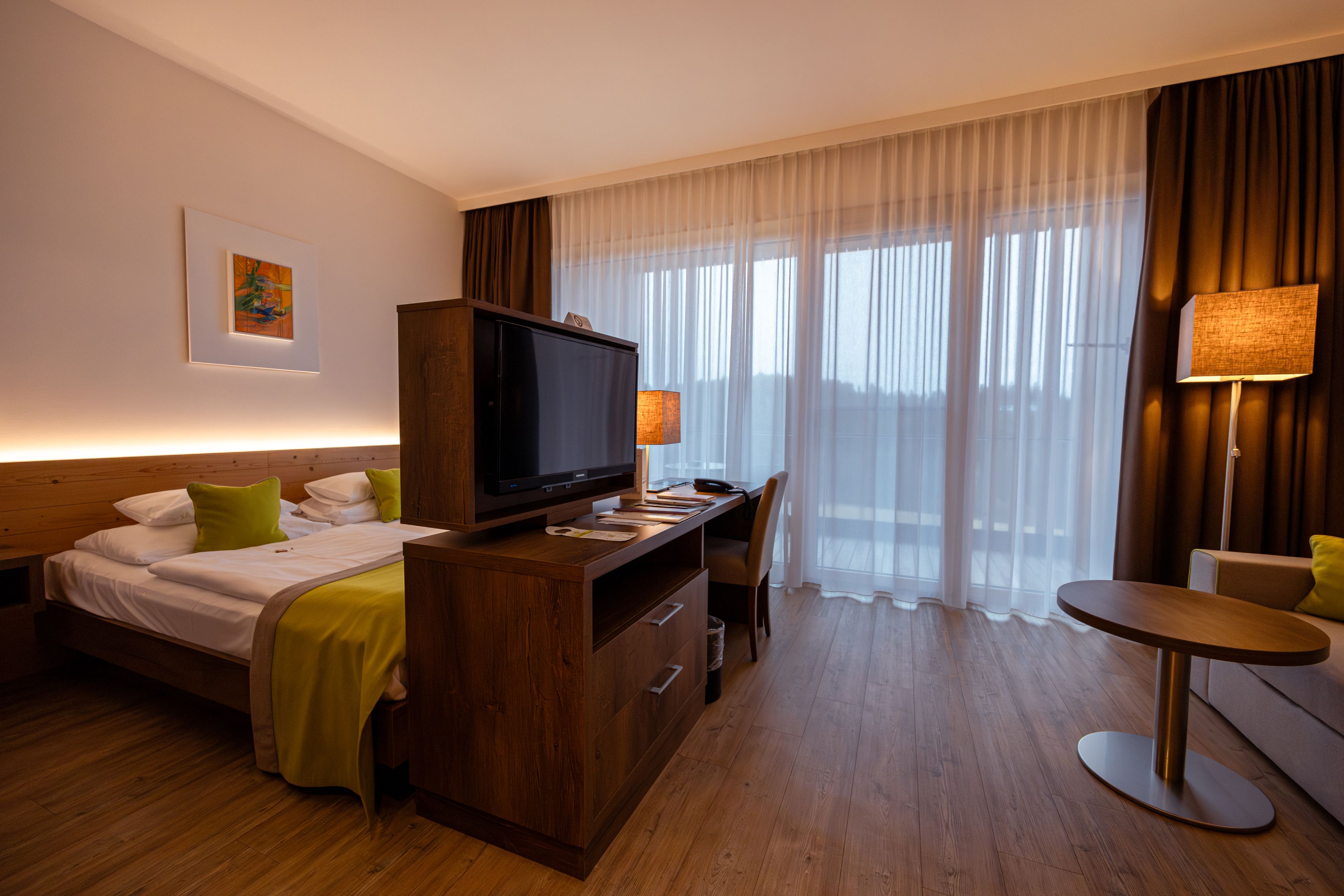 Modernes Hotelzimmer mit Doppelbett, TV, Schreibtisch und großen Fenstern mit Vorhängen.