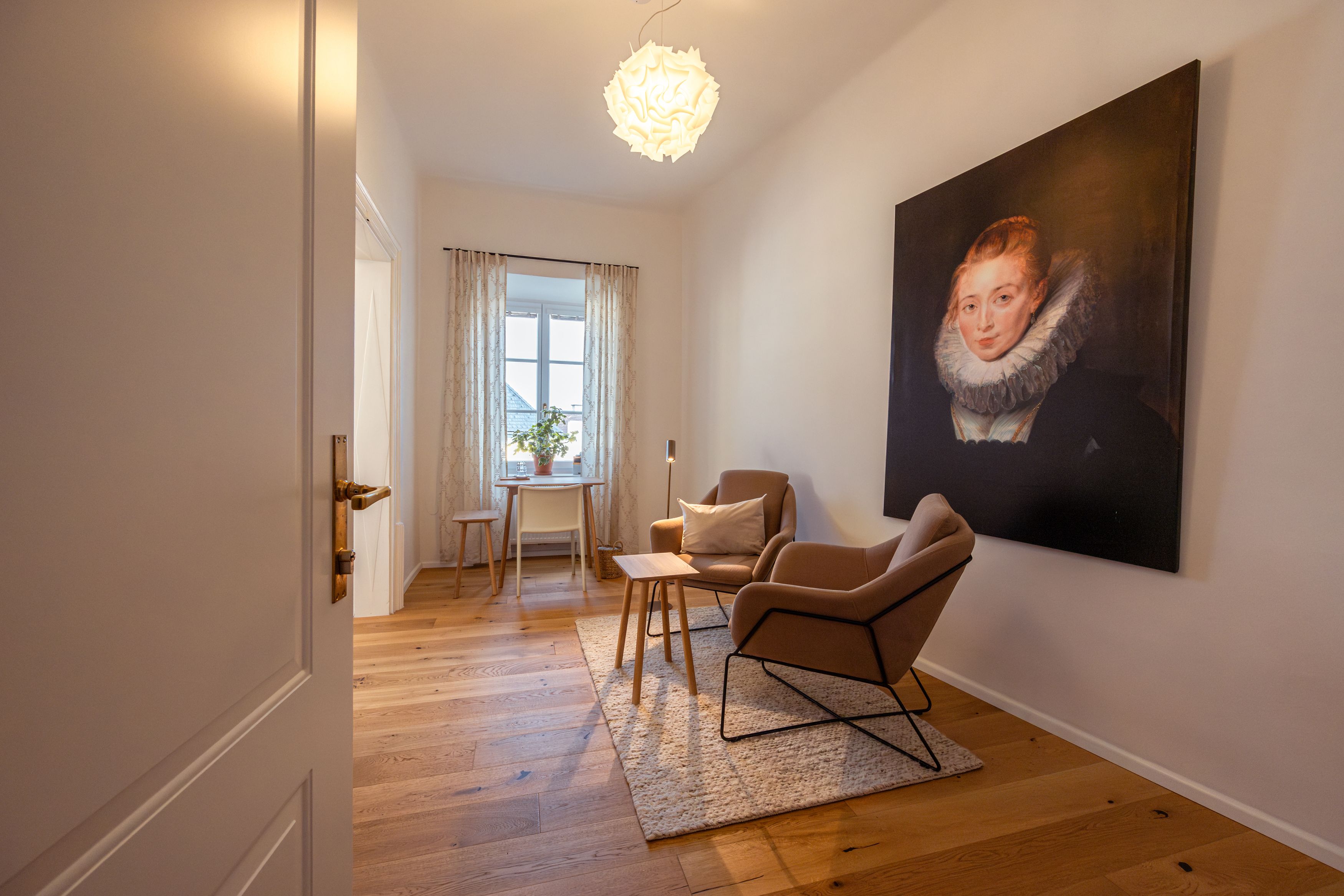 Ein gemütlicher Raum mit zwei Sesseln, einem Tisch und einem großen Porträt an der Wand. Ein Fenster mit Vorhängen lässt Tageslicht herein.