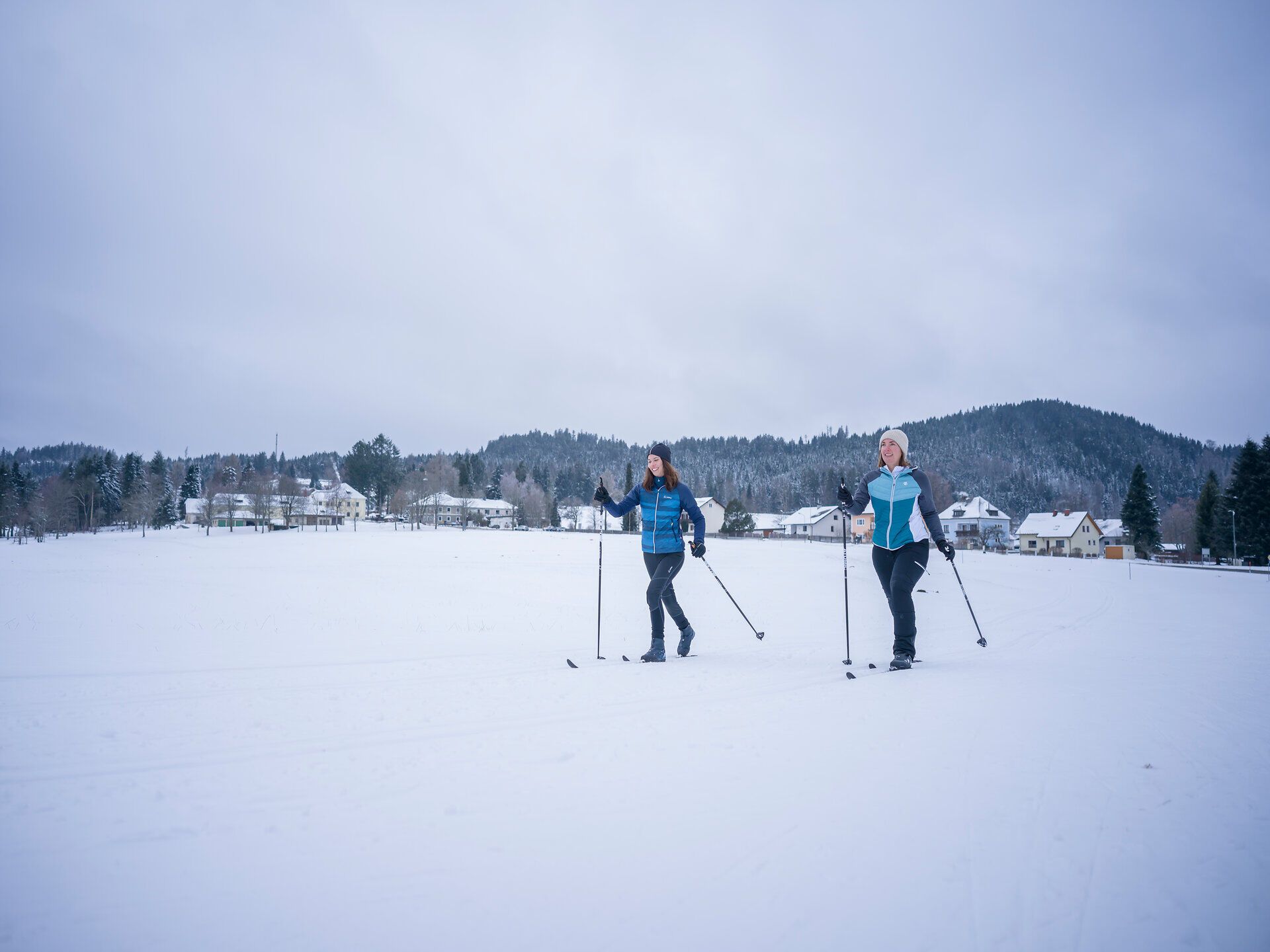 Zwei Personen beim Langlaufen auf einer schneebedeckten Fläche vor einem Dorf und bewaldeten Hügeln.