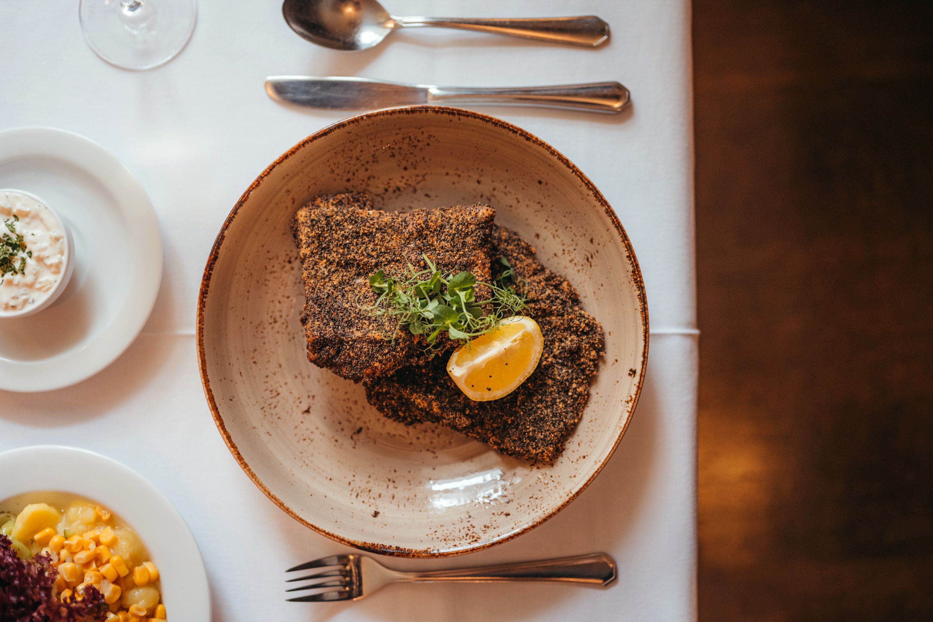 Graumohn-paniertes Karpfenfilet mit Zitronenscheibe und Kräutern auf Teller, daneben Beilagen.