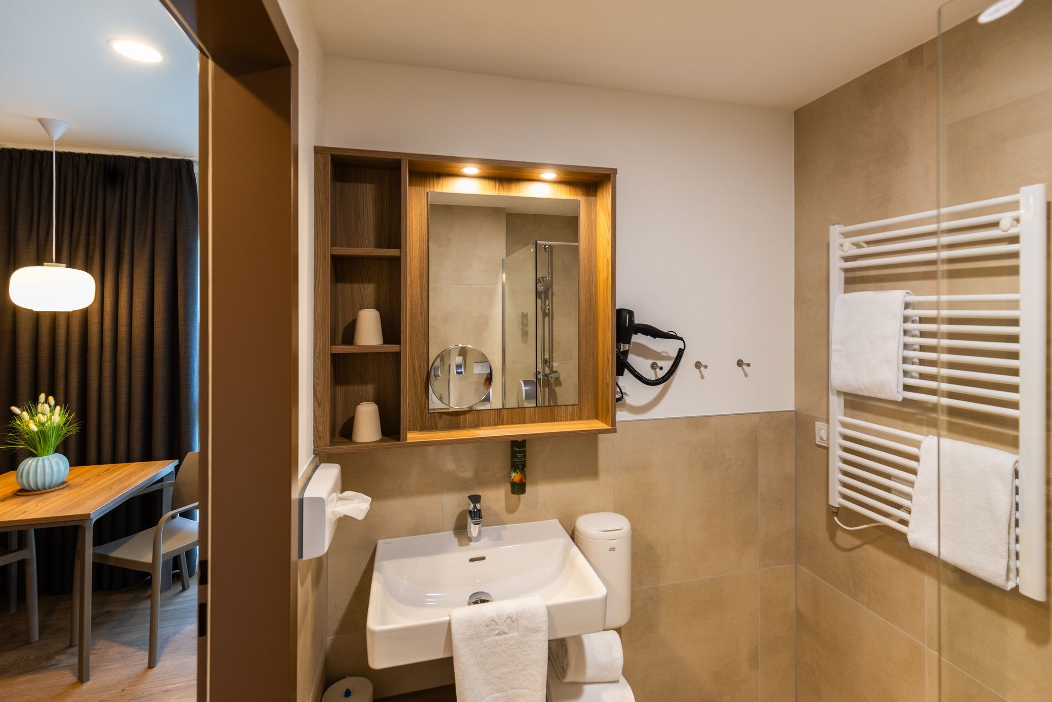 Modernes Badezimmer mit Waschbecken, Spiegel, Handtuchhalter und Blick in einen angrenzenden Raum mit Tisch und Vase.