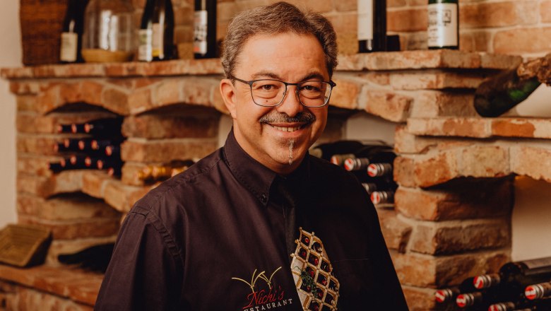 Ein Mann mit Brille und Schnurrbart steht l&auml;chelnd vor einem Weinregal in einem Restaurant.