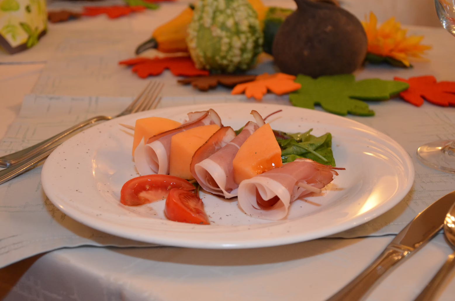 Ein Teller mit Schinken und Melone, dekoriert mit Tomaten und Salat, auf einem Tisch mit herbstlicher Dekoration.