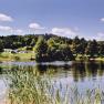 Ein idyllischer Campingplatz mit Zelten am Ufer eines Sees, umgeben von B&auml;umen und Wiesen unter blauem Himmel.