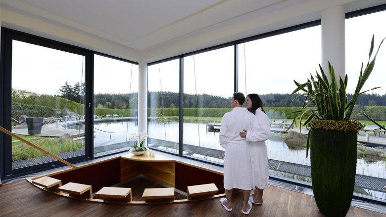 Ein Paar in Badem&auml;nteln steht in einem modernen Wellnessbereich mit Blick auf einen Teich.