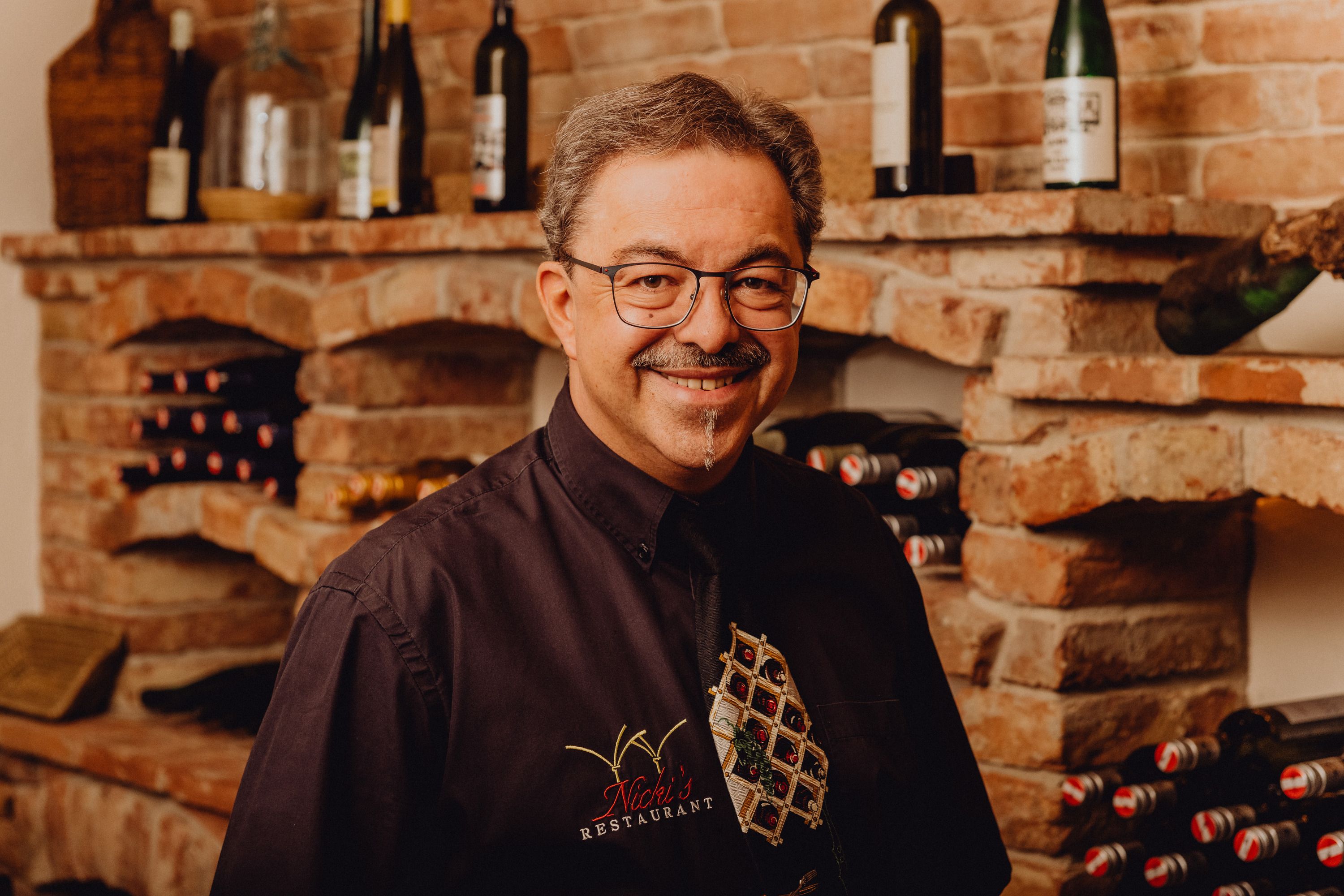 Ein Mann mit Brille und Schnurrbart steht lächelnd vor einem Weinregal in einem Restaurant.