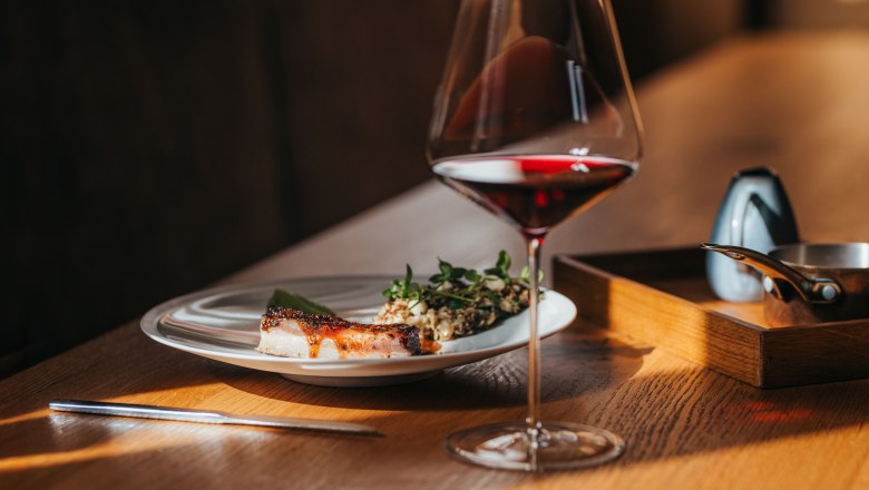 Ein Teller mit Lammbauch, Kopfsalat und Wasabicr&egrave;me neben einem Glas Rotwein auf einem Holztisch.