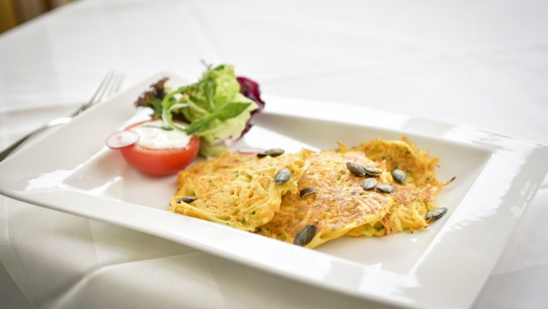 Ein Teller mit Kartoffelpuffer, Kürbiskernen und Salat auf einem weißen Tisch.