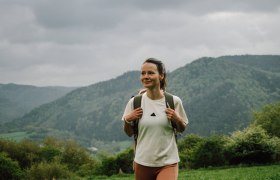 Ein Wanderer genie&szlig;t die frische Bergluft und die atemberaubende Aussicht auf die sanften H&uuml;gel und dichten W&auml;lder. Die Wolken am Himmel verleihen der Szenerie eine mystische Atmosph&auml;re, w&auml;hrend die Natur in voller Bl&uuml;te steht. Diese Idylle l&auml;dt dazu ein, die Seele baumeln zu lassen und die Sch&ouml;nheit der Umgebung zu erkunden.