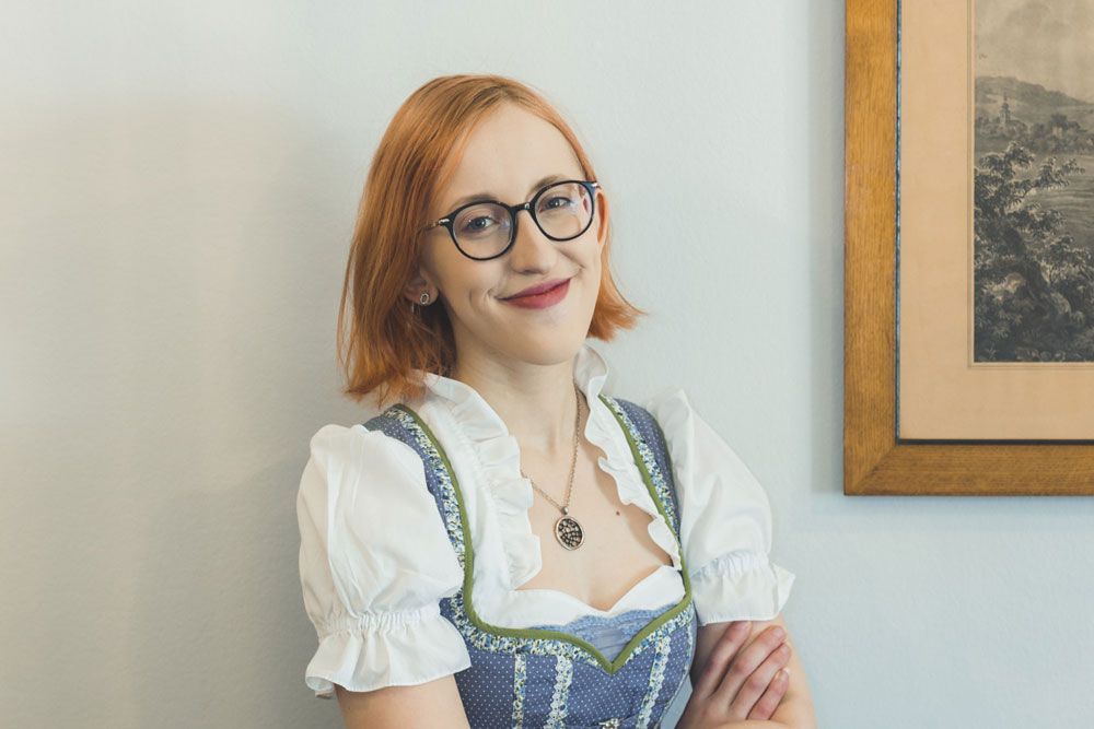 Eine Frau mit Brille und Dirndl lächelt vor einem Bild an der Wand.
