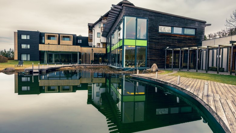 Modernes Wellnesshotel mit Holzfassade und gro&szlig;em Teich im Vordergrund.