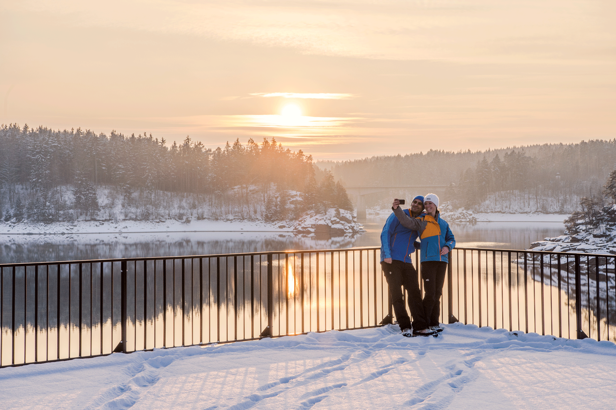 Zwei Personen machen ein Selfie am verschneiten Stausee Ottenstein bei Sonnenuntergang.