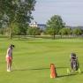 Golferin mit Hund auf gr&uuml;nem Golfplatz, B&auml;ume und Geb&auml;ude im Hintergrund.