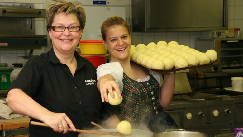 Zwei Frauen in einer K&uuml;che, eine h&auml;lt einen Tablett mit Kn&ouml;deln, die andere kocht Kn&ouml;del in einem Topf.