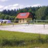 Menschen spielen Volleyball auf einem Sandplatz vor einem gelben Geb&auml;ude mit rotem Dach, umgeben von B&auml;umen.