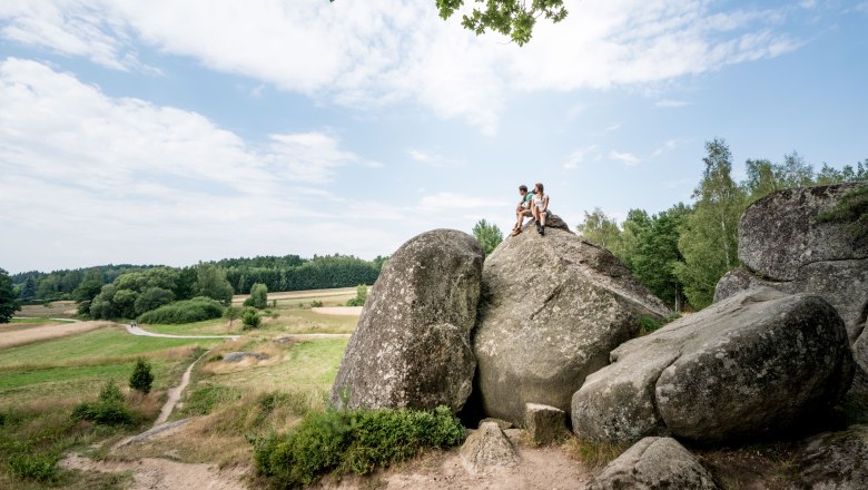 Zwei Personen sitzen auf einem gro&szlig;en Felsen in einer l&auml;ndlichen Landschaft mit Wiesen und B&auml;umen.