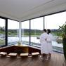 Ein Paar in Badem&auml;nteln steht in einem modernen Wellnessbereich mit Blick auf einen Teich.