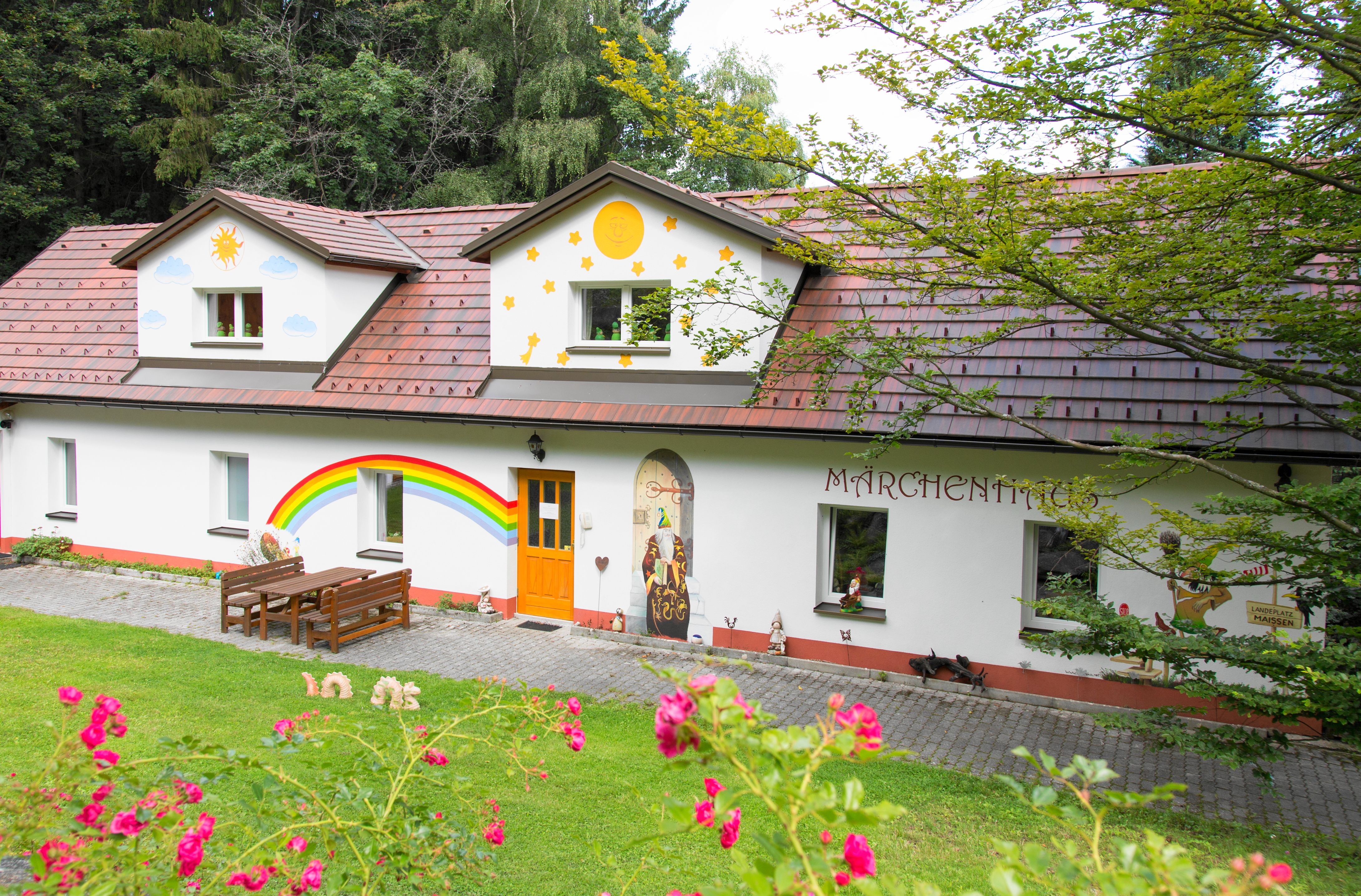 Ein bunt bemaltes Haus mit Märchenmotiven und einem Regenbogen an der Wand, umgeben von grüner Natur.
