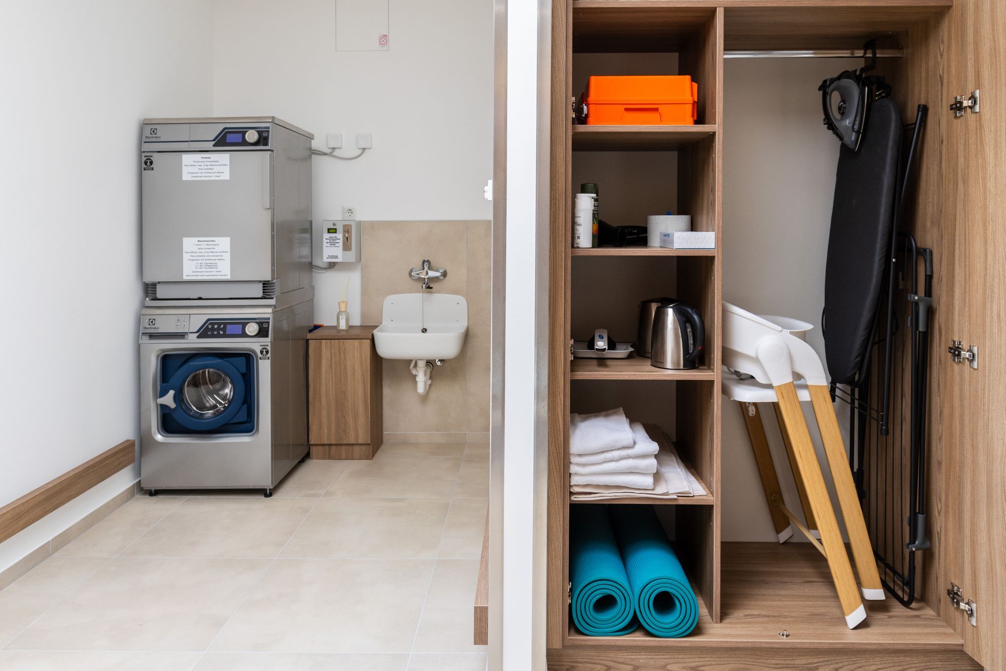 Ein Waschraum mit Waschmaschine, Trockner, Waschbecken und einem Schrank mit Haushaltsgegenständen wie Bügelbrett und Wasserkocher.