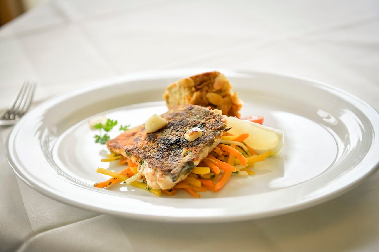 Ein Teller mit gebratenem Fischfilet auf Gemüsestreifen und einer Beilage auf weißem Tischtuch.
