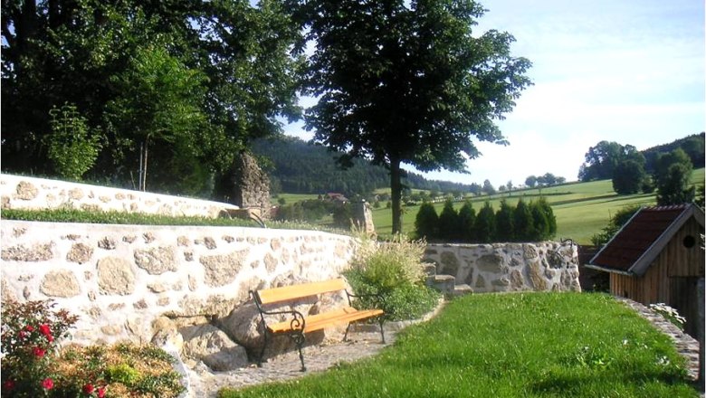 Ein Garten mit Steinmauer, Bank und B&auml;umen im Hintergrund.